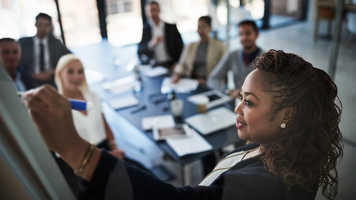 Woman writing on whiteboard in front of staff surrounding board room table